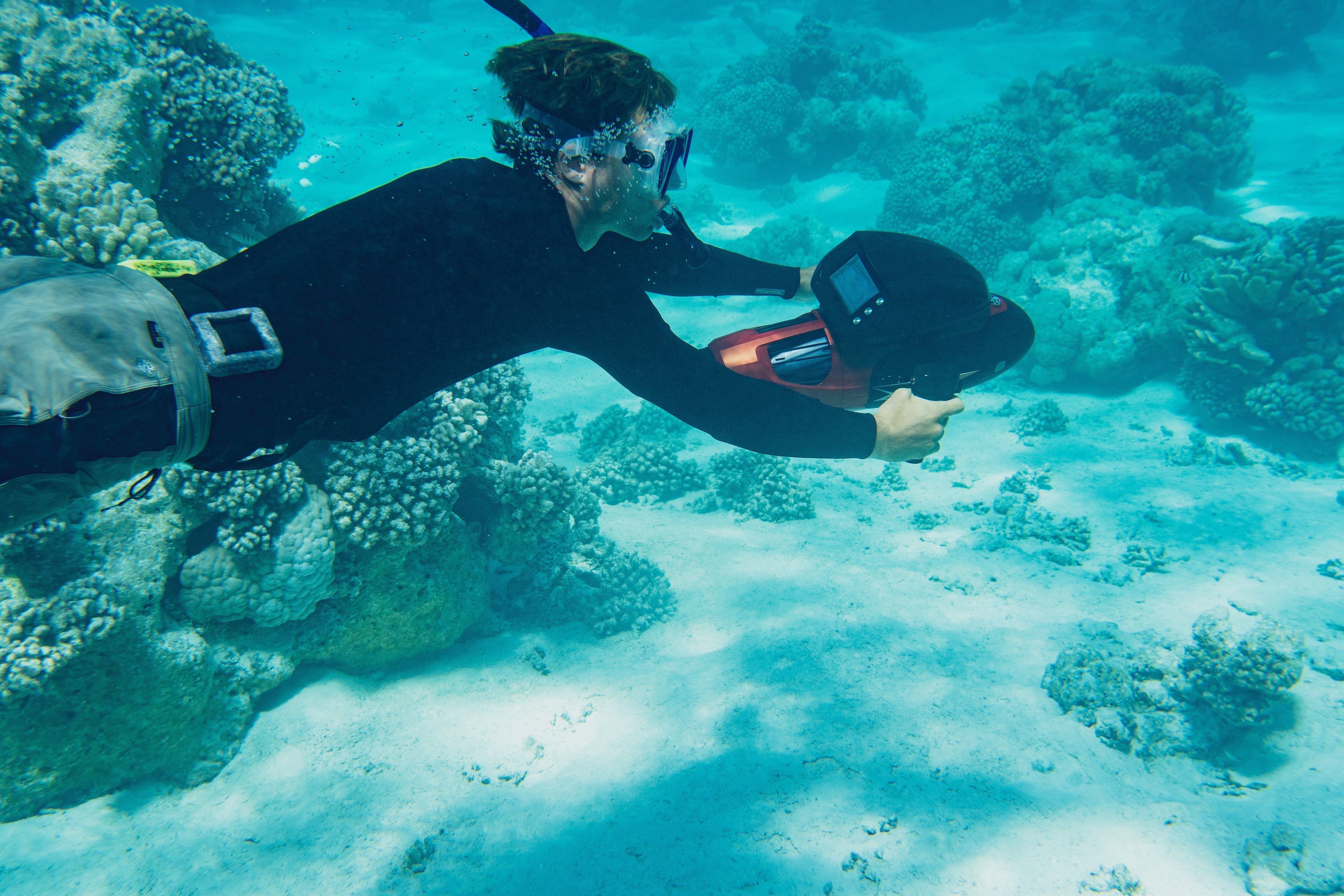 A diver in a wetsuit swims near coral reefs, holding an underwater scooter, with clear blue waters surrounding them.