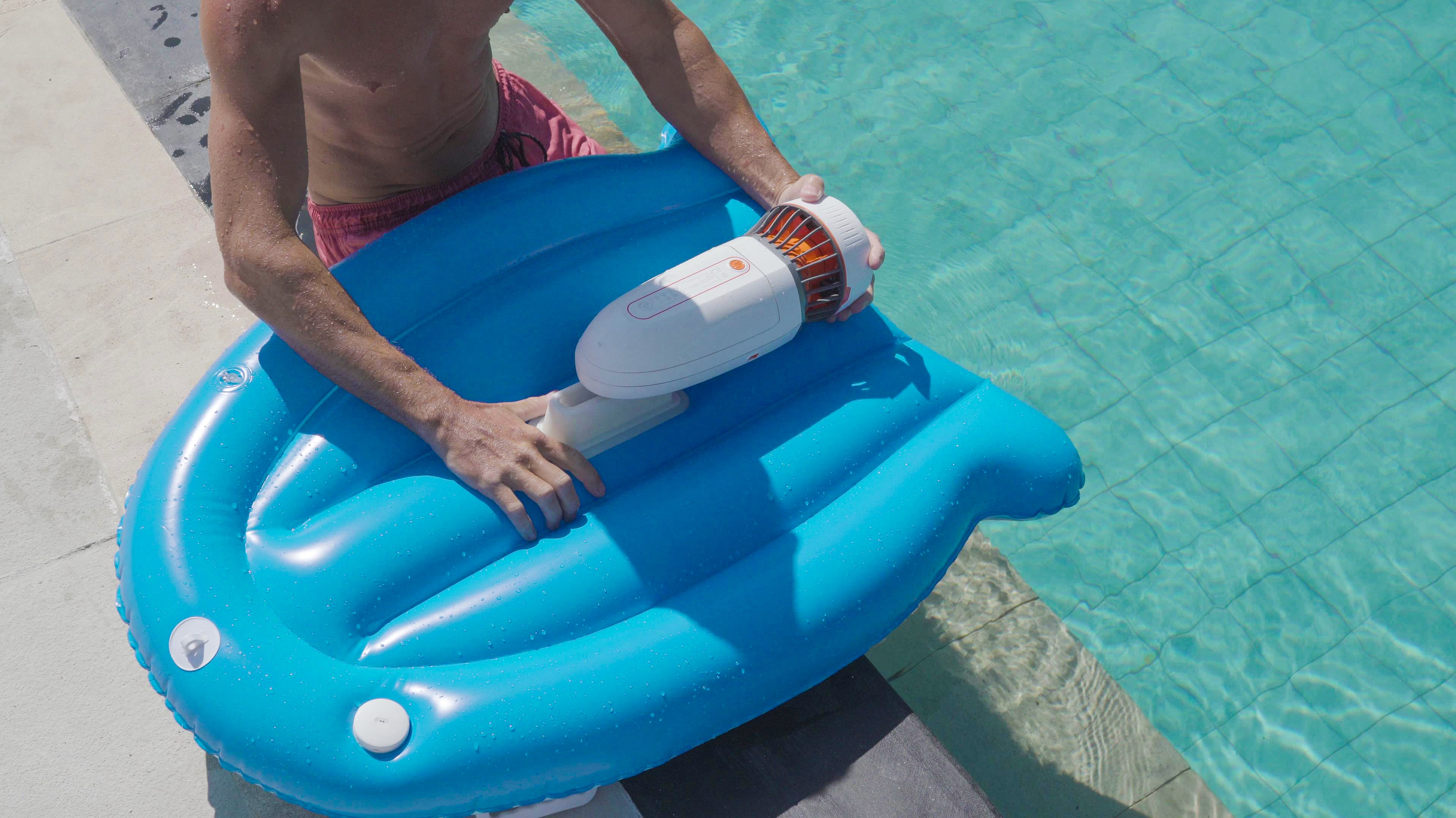 User holding Tini underwater scooter on blue kickboard in sunny pool.