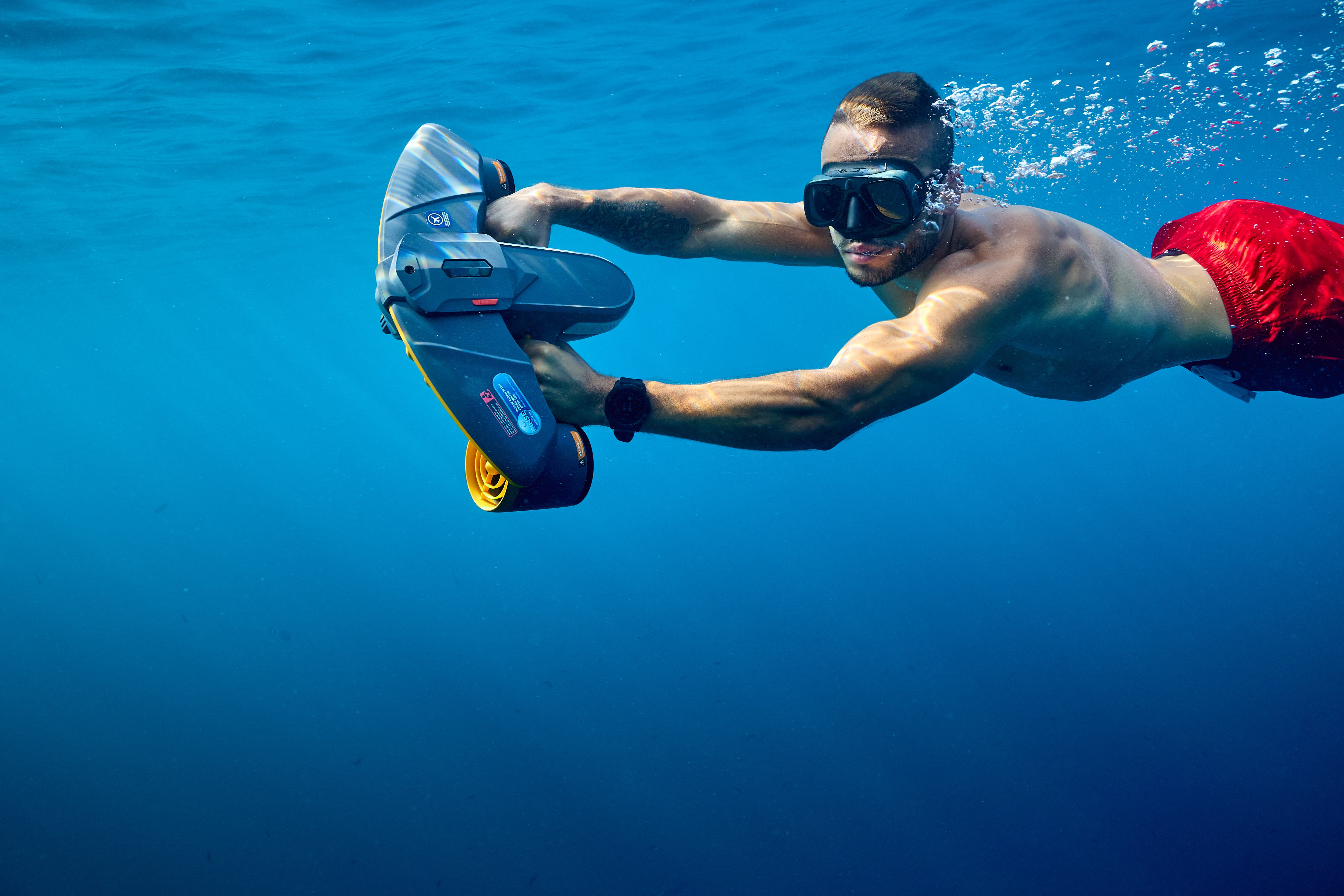 Diver in red shorts using SUBLUE Navbow+ underwater scooter in clear blue water.