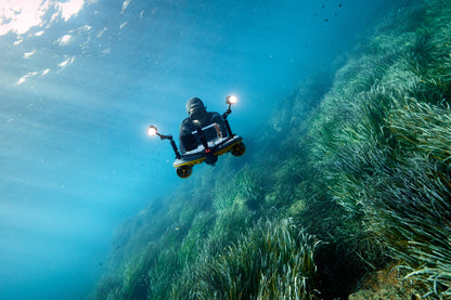 Diver on Sublue scooter exploring underwater seagrass with twin LED lights.