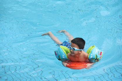 Child swimming with parrot armbands and orange swim board in a blue pool.