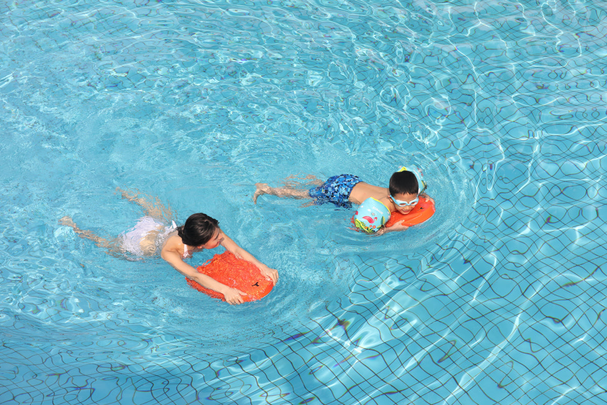 Two kids swimming with an orange swim board and beach ball in blue pool