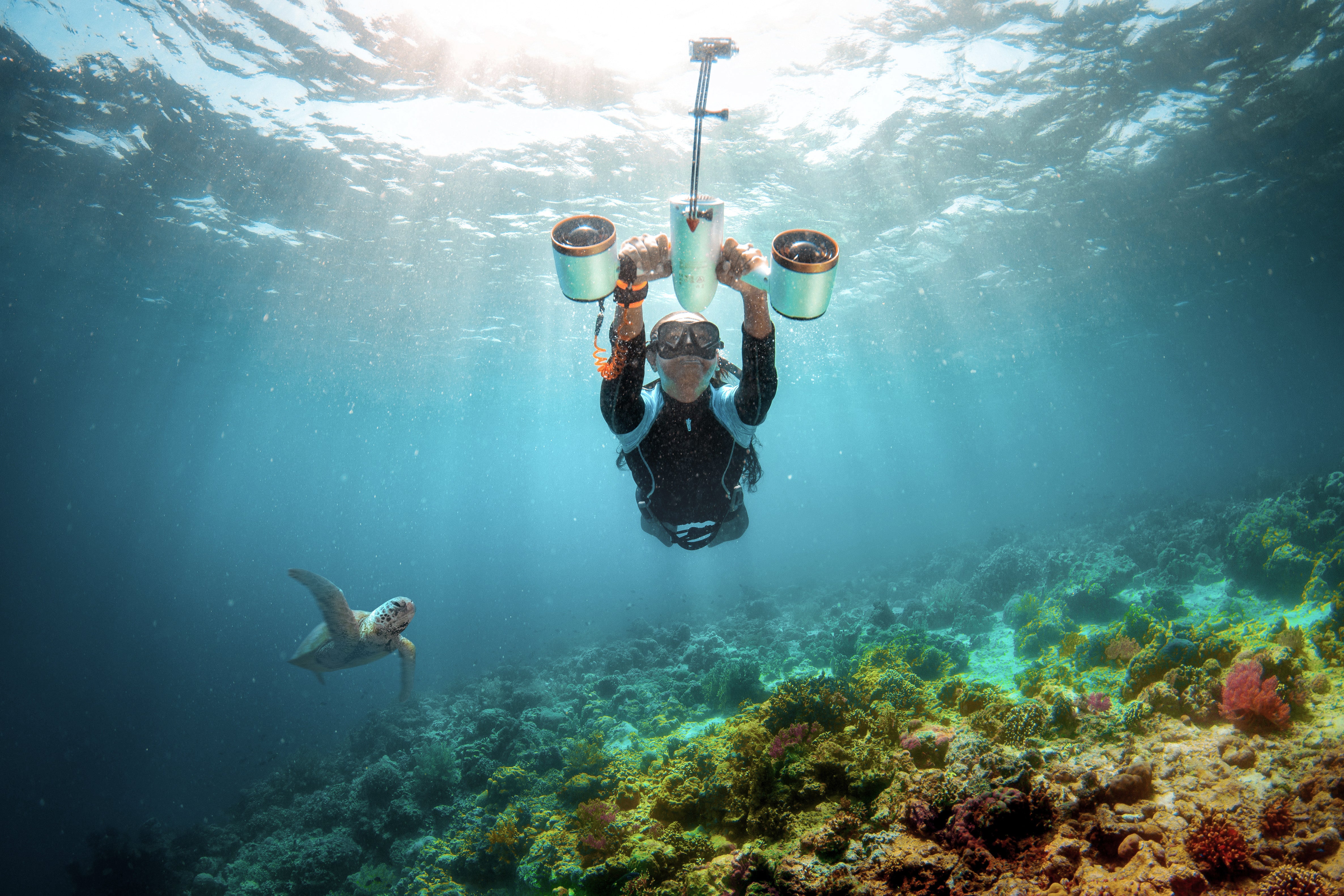 Diver with Sublue scooter and selfie extension stick near coral reef and sea turtle.